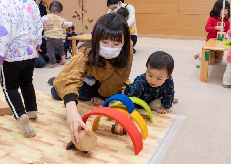 青や黄色、赤などの小さなトンネルのようなものの下を木製のおもちゃを押して遊ぶ子供と母親の写真