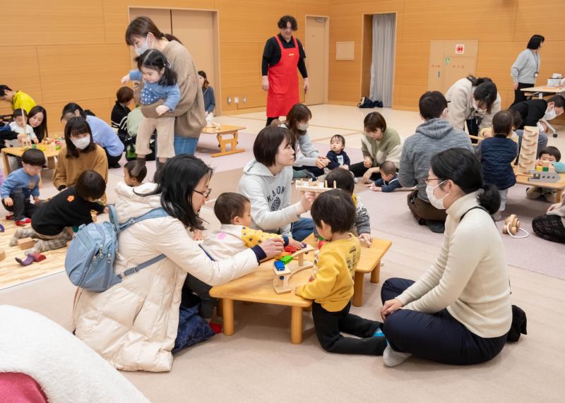 交流センター内に置かれた複数のテーブルの上で木製のおもちゃで遊ぶたくさんの親子の写真