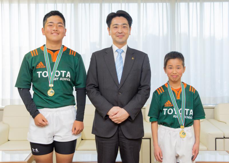 緑色のユニフォーム姿で首にメダルをかけた西浦さん、山田さんが市長を挟み記念撮影をしている写真