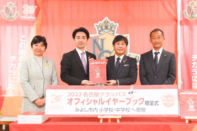 贈呈式で市長と清水克洋専務取締役が名古屋グランパスオフィシャルイヤーブックを2人で持ち、関係者男女1名ずつ隣に並び、4名で記念撮影をしている写真