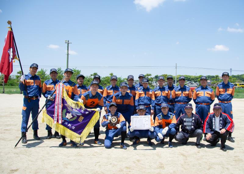大会で優勝した莇生分団が3列に分かれて整列し、優勝旗、賞状を広げて撮影された集合写真