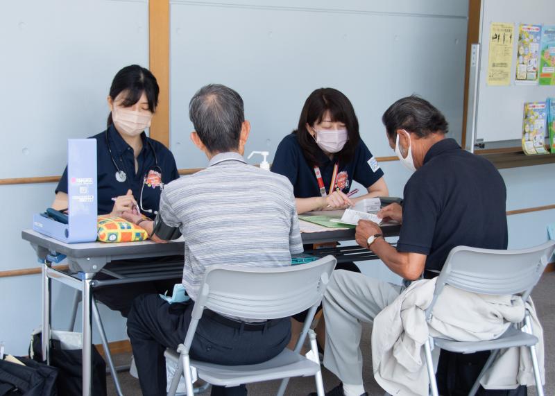 健康相談コーナーで1人の男性は保健師に血圧を測定してもらい、もう1人の男性は別の保健師に健康状態を相談している写真