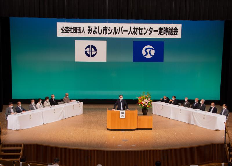 公益社団法人みよし市シルバー人材センター定期総会と書かれた幕のステージに座っている来賓の前で議長があいさつをしている写真