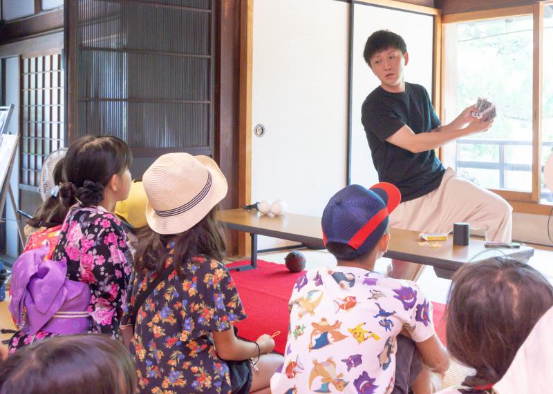 男性によるカードマジックのパフォーマンスを見ている子供達の後ろ姿を写した写真
