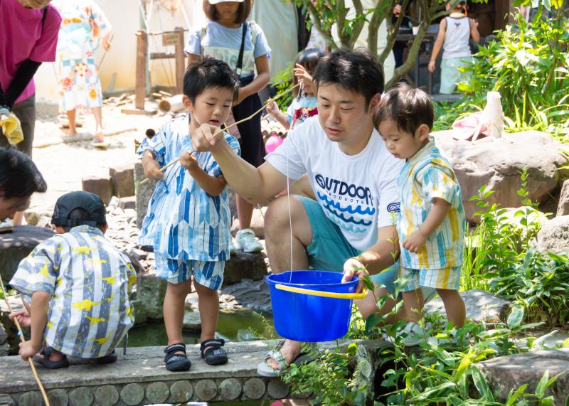 紐の付いた竿を男の子とお父さんが一緒に持ち、紐の先端を青いバケツの中に入れているザリガニ釣り大会の写真
