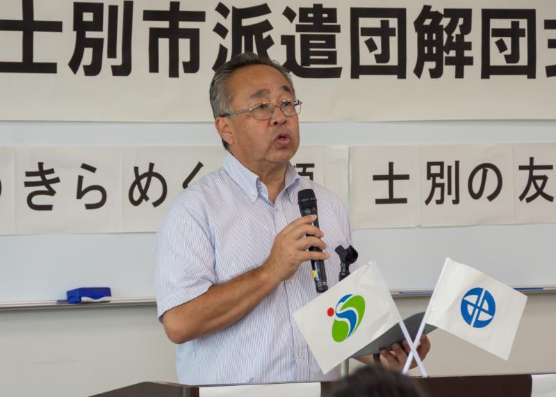 演壇に北海道士別市とみよし市の市章の小さい旗が飾られており、男性がマイクを持ち話している写真