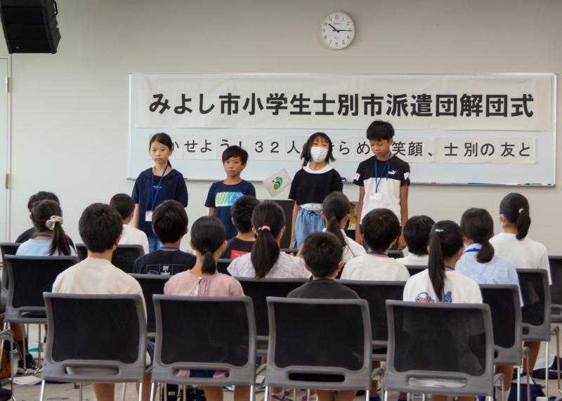 「みよし市小学生士別市派遣団解団式」と書かれた横長の紙がホワイトボードに貼られ、その前に4名の児童がたっている写真