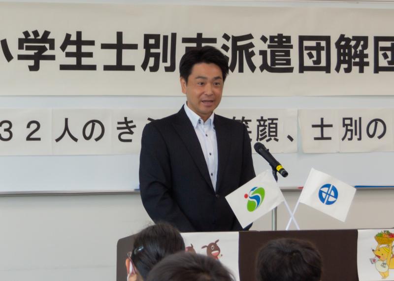 演壇に北海道士別市とみよし市の市章の小さい旗が飾られており、小山祐市長が演壇で話している写真