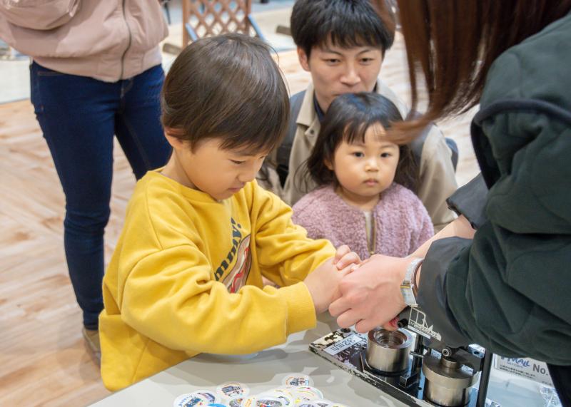 黄色い洋服を着た男の子が、女性の方と一緒に缶バッジ作りをしている様子の写真