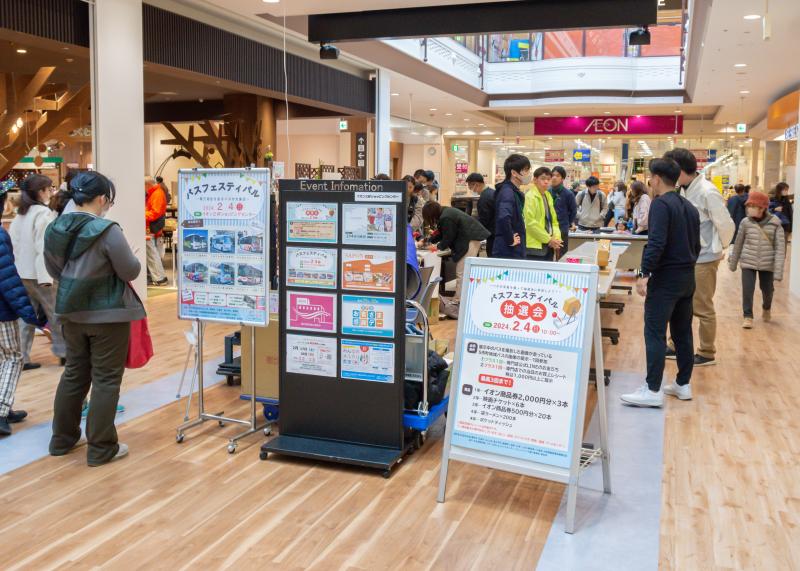 「バスフェスティバル」や「バスフェスティバル抽選会」と書かれたポスターが貼られたボードがショッピングセンターの通路に置かれ、催しが行われている様子の写真