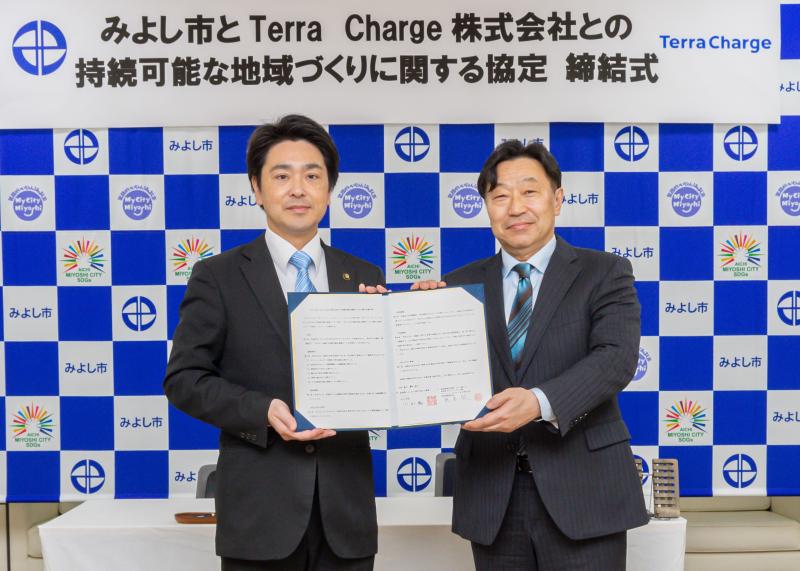 みよし市とTerra Charge株式会社との接続可能な地域づくりに関する協定 締結式の幕の前で本田さんと市長が締結書を持ちながら記念撮影をしている写真