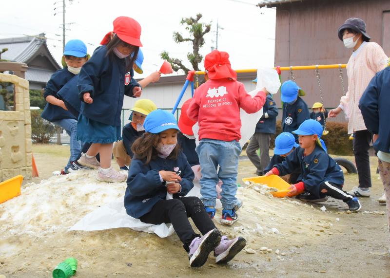 山のように積まれた雪の上を女の子がビニール袋を使ったそり滑りをしている写真