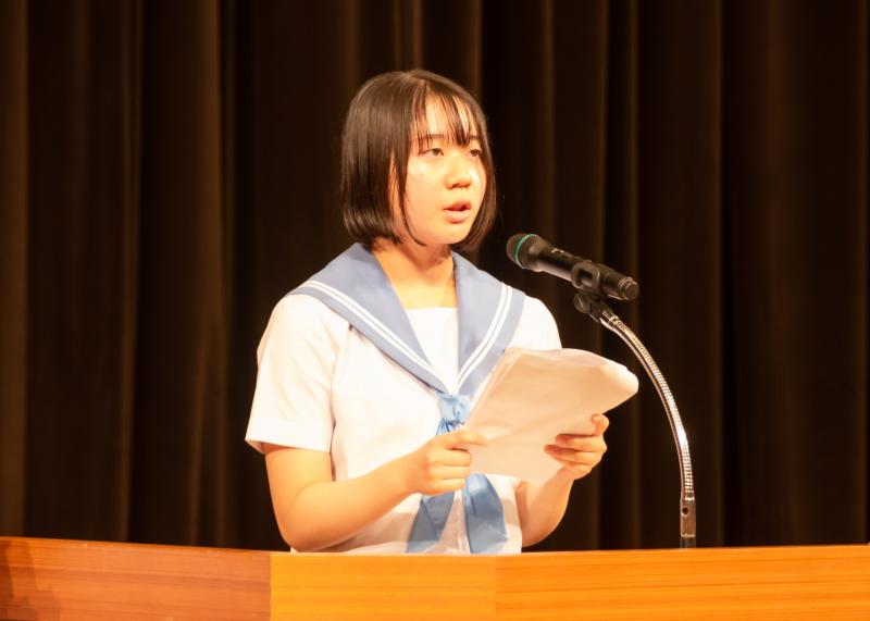 水色の襟のセーラー服姿の女子生徒が壇上のマイクの前に立ち「みよし市中学生平和学習広島派遣報告」をしている様子をアップで撮影した写真