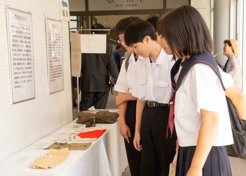 制服姿の2人の男子生徒と女子生徒が展示物の前に足をとめ資料を見ている様子を撮影した写真