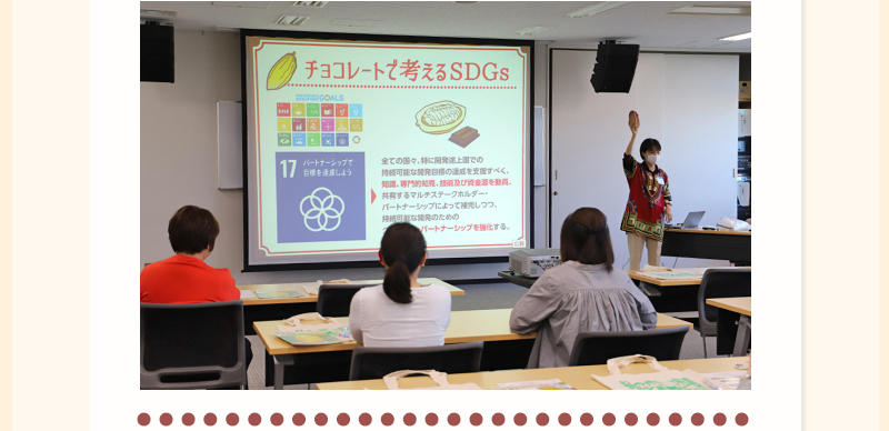 「チョコレートで考えるSDGs」と書かれたスクリーンの横で稲垣さんが手に持ったカカオを上にあげながら話をしている写真