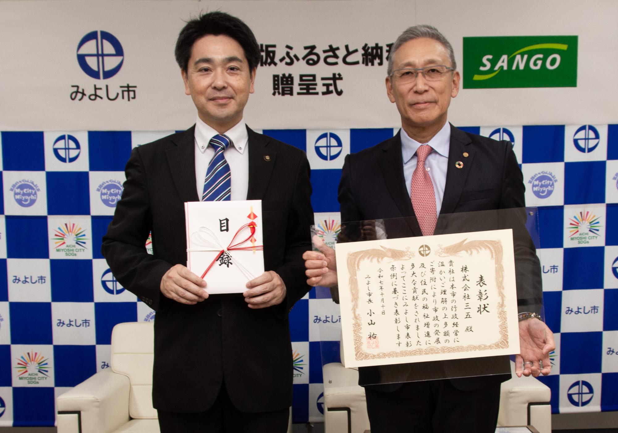 みよし市のバックボードの前で市長と水野代表取締役社長が表彰状を持ち記念撮影をしている写真