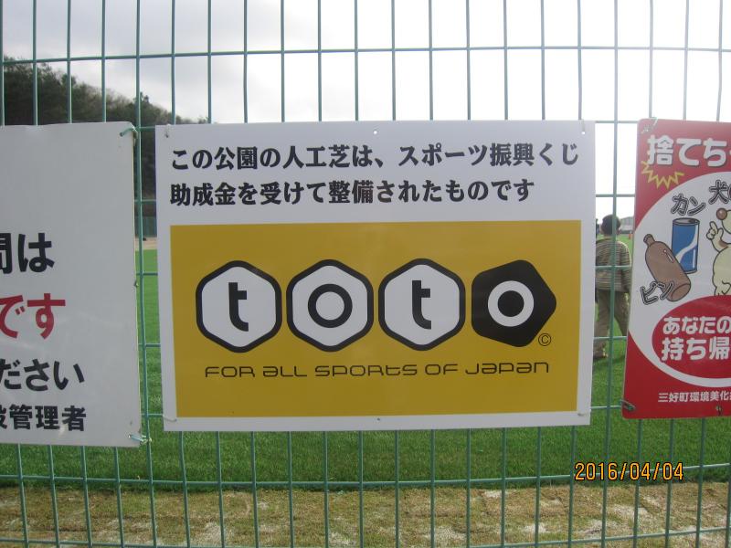 公園のフェンスにこの公園の人工芝は、スポーツ振興くじ 助成金を受けて整備されたものですと書かれ黄色の看板にtotoと書かれた看板の写真