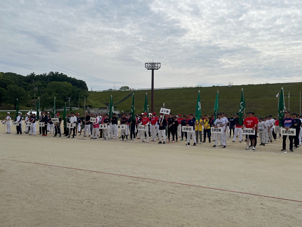 グラウンドにユニフォームを着た選手たちが、地区のプラカードを持っている方を先頭に整列している様子