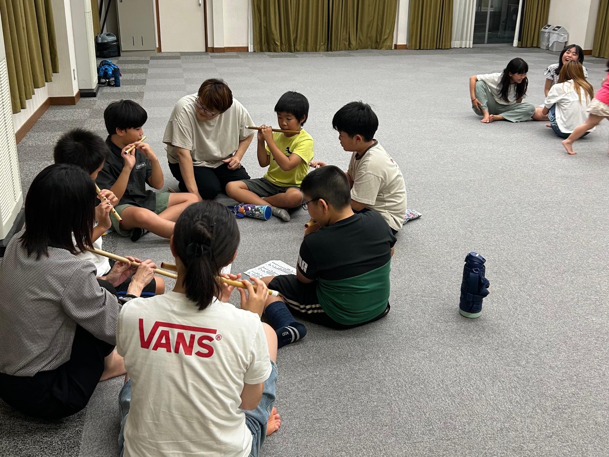 室内で輪になって座った子どもたちが大人に見守られながら横笛を演奏している写真