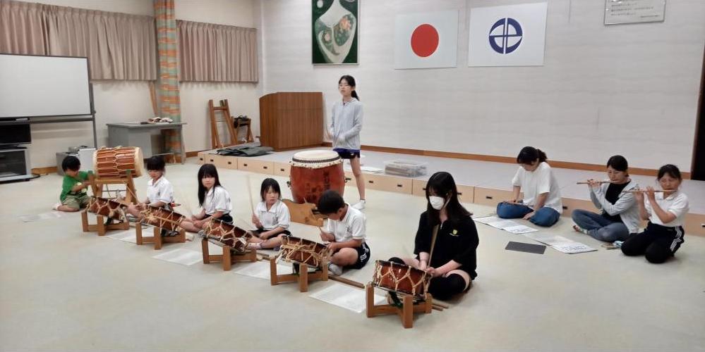 室内の舞台の前で太鼓や笛の練習をしている子どもたちの写真
