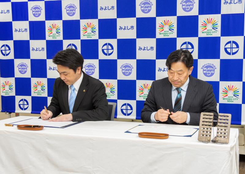 みよし市のバックボードの前に白いクロスが敷かれた長テーブルを置きその前に着席した市長とTerra Charge株式会社の関係者が締結書にサインをしている様子の写真