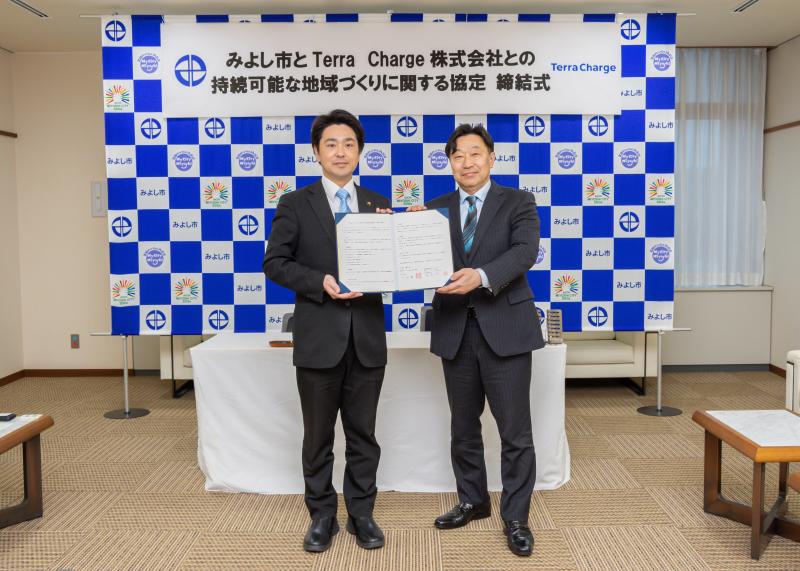 「みよし市とTerra Charge株式会社との持続可能な地域づくりに関する協定 締結式」と書かれた横断幕とバックボードの前で締結書を一緒に持った市長とTerra Charge株式会社の関係者が記念撮影をしている写真