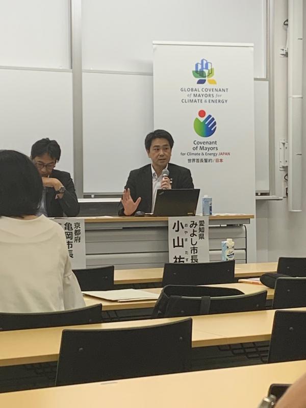 前に座る亀岡市長とマイクを持って話すみよし市長を撮影した写真