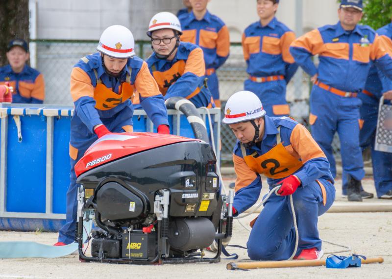 機械の後ろで立って操作する消防団員の男性と、機械の横で片膝をつき操作する消防団員の男性の写真