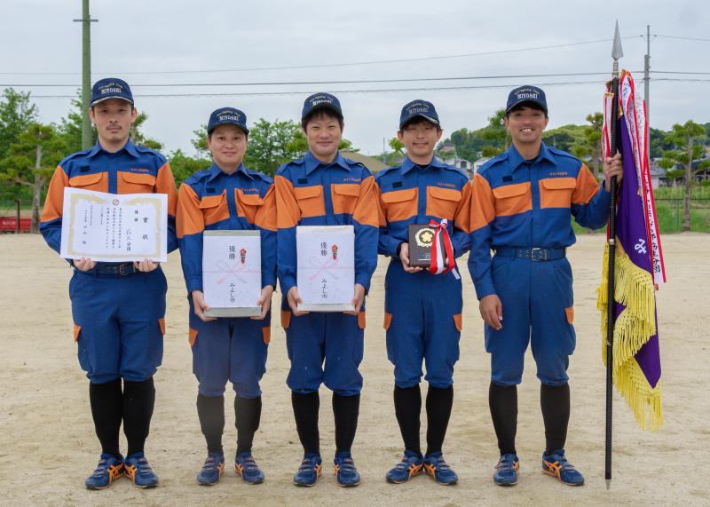 左から賞状、「優勝」とかかれた熨斗が貼られた記念品、表彰盾、優勝旗をそれぞれ持つ5名の消防団員が並んだ写真
