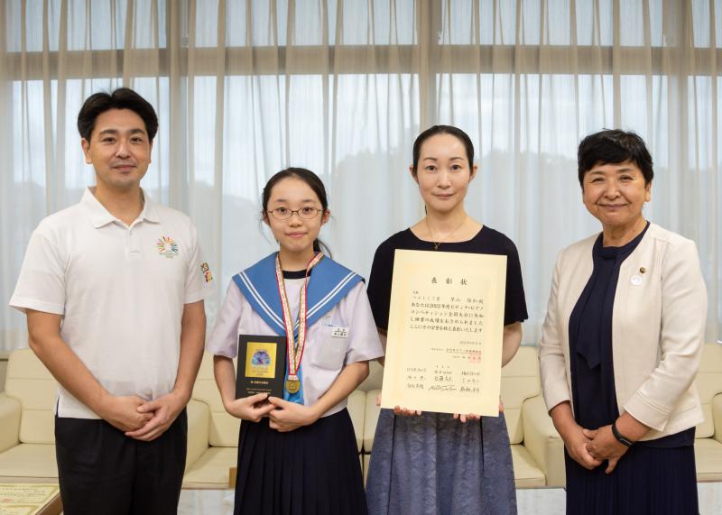 右から市長、北中学校2年生の草山陽和さんがメダルを首にかけ盾を持ち、賞状を持った女性、白いジャケットを着た女性の4人で1列に並び報告会で記念撮影をしている写真