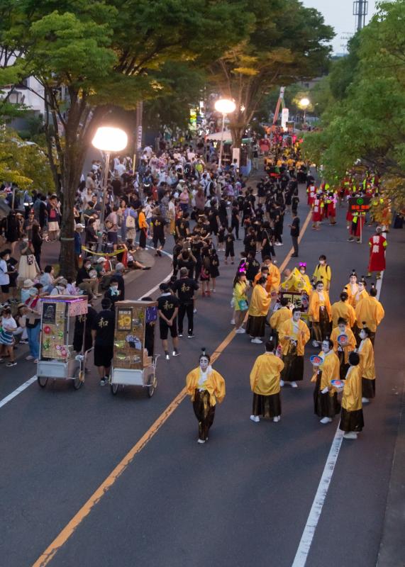 道路の左側には和と書いた黒のTシャツ、黒の短パン姿のグループ、右には上は黄色、下は暗めの金色の袴を着てちょんまげをし顔は真っ白なバカ殿さま風の姿をしたグループが立って待っているような写真