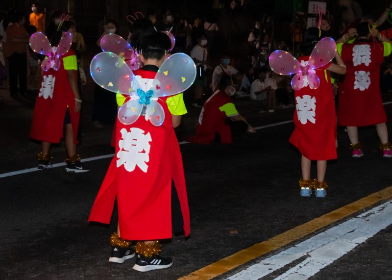 赤の法被を着て背中にイルミネーションが付いた蝶々の羽を付け踊るグループの写真