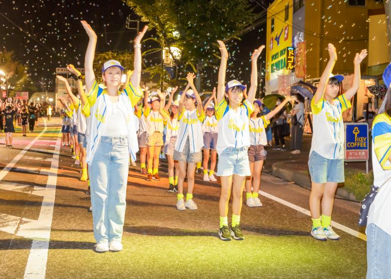白を基調とし袖の所が黄色のシャツ、帽子の庇が紫色の衣装姿のグループが踊りシャボン玉が飛んでいる写真