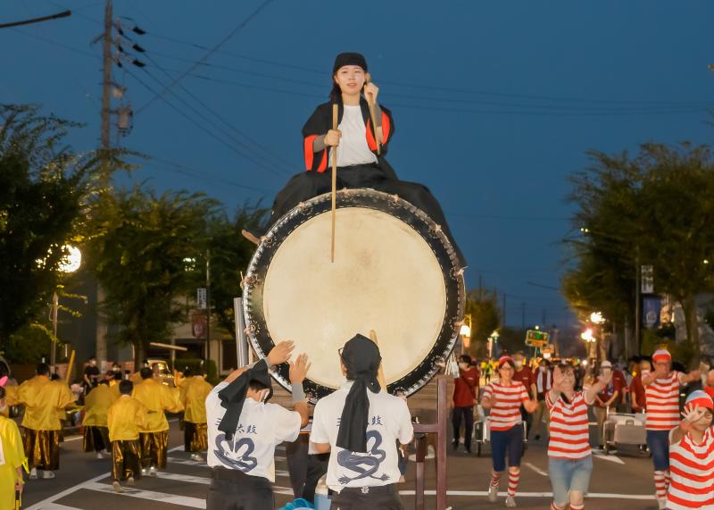 大きな太鼓の上に座りバチで叩きながら行進している写真